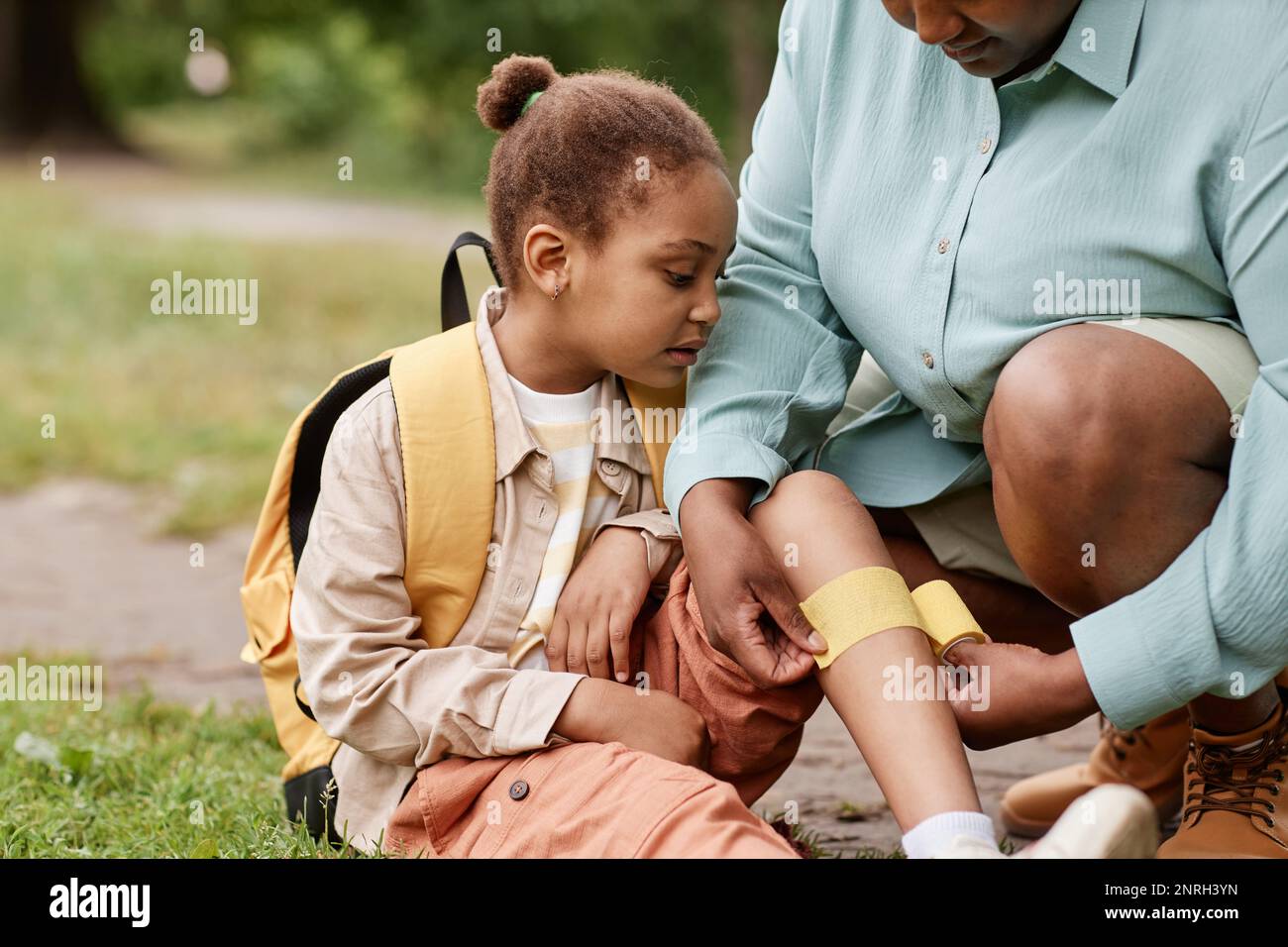 Portrait of black little girl injured in nature hike with mother putting bandage on knee outdoors Stock Photo