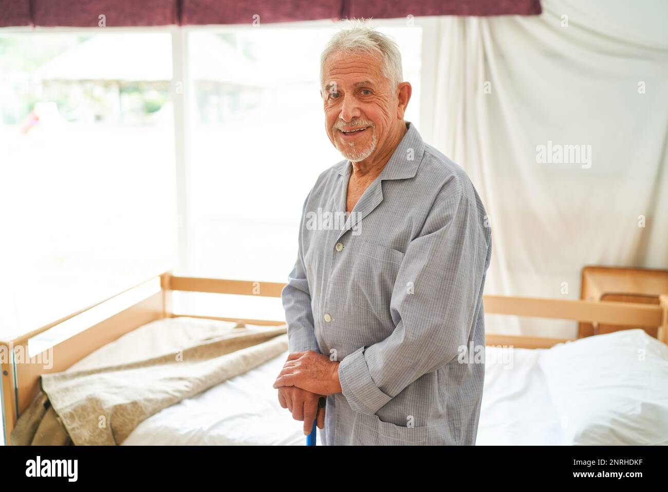 Portrait of smiling senior man holding walking cane standing by bed at retirement home Stock Photo
