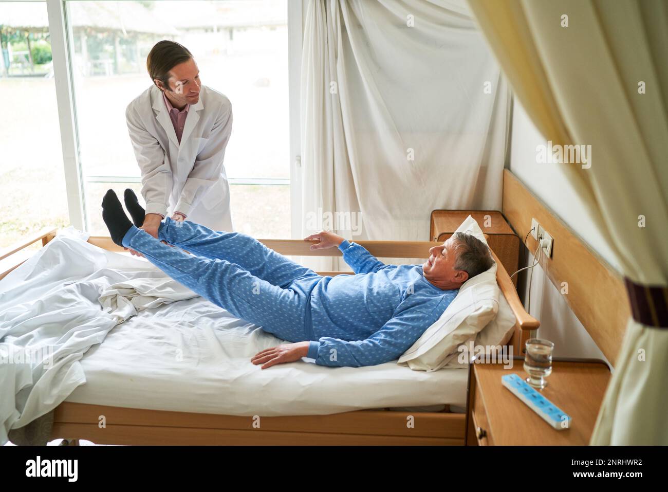 Smiling male doctor lifting legs of senior man lying in bed at