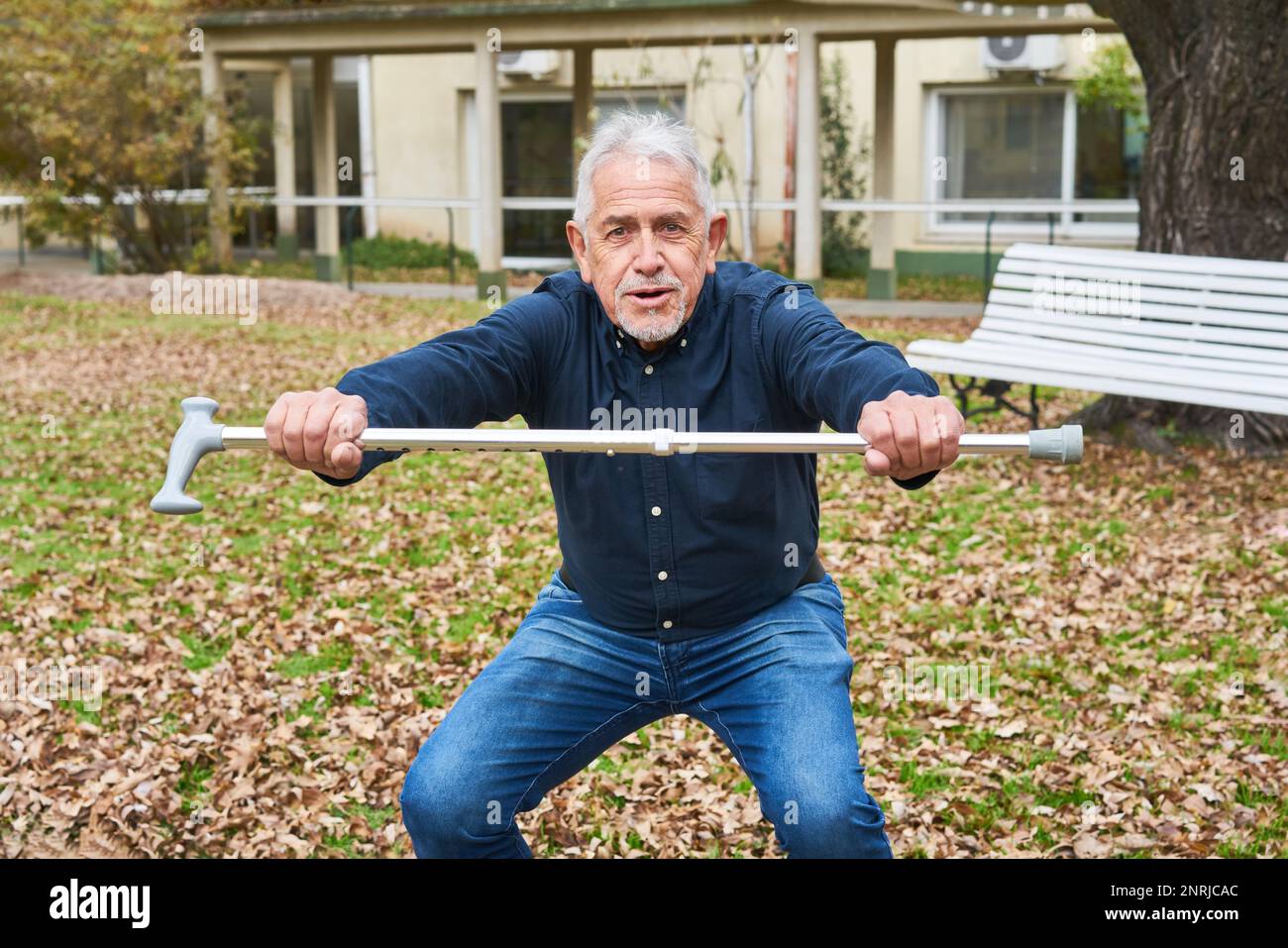 Portrait of elderly man squatting while holding walking cane in garden of nursing home during autumn Stock Photo