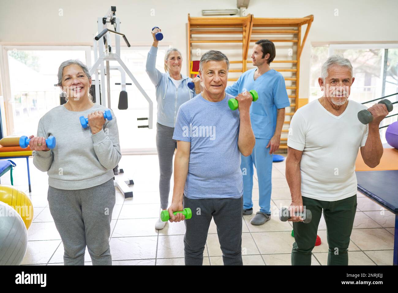 Elderly men and woman exercising with dumbbells while therapist assisting patient in background at nursing home Stock Photo