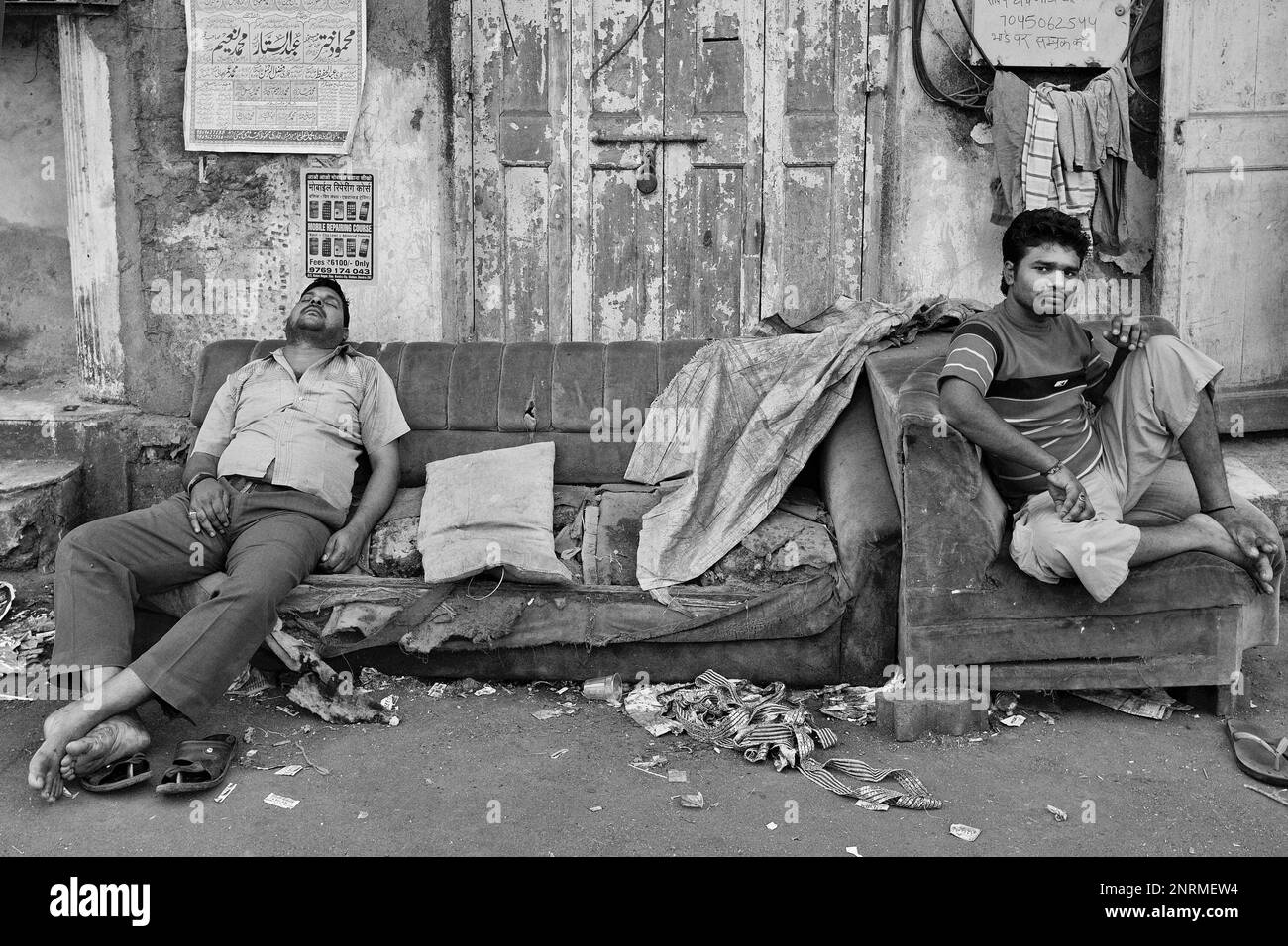 An Indian man sleeping on a discarded, dilapidated sofa, another man sitting on a junked armchair; outside in Arab Galli, Grant Rd., Mumbai, India Stock Photo