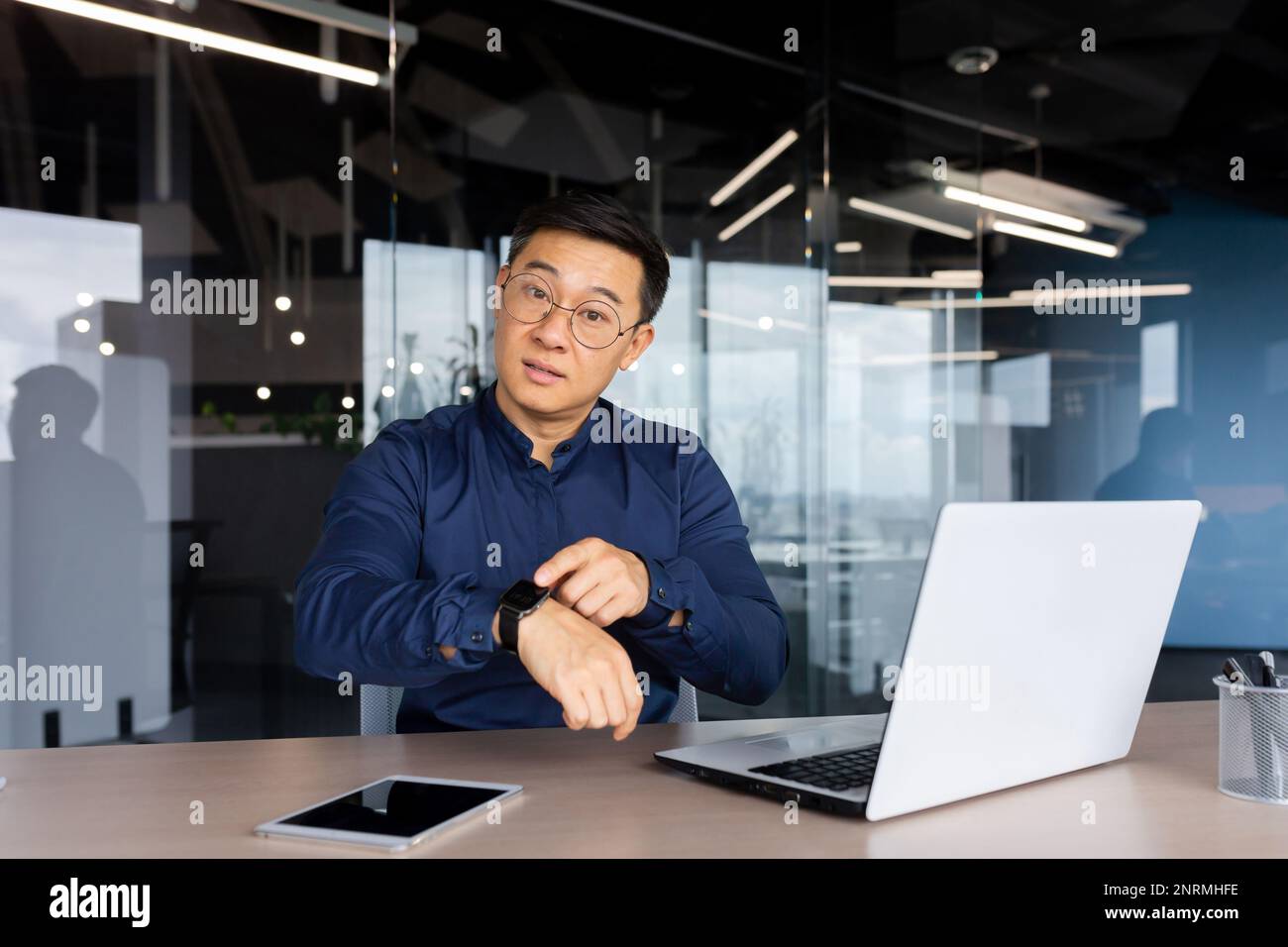 Man at work with laptop inside office, businessman unhappy and serious showing watch to camera, being late for colleague's meeting. Stock Photo