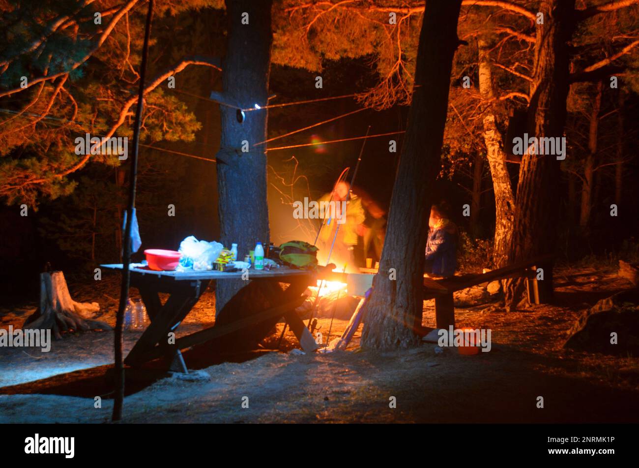 A tent stands at night by a fire with a table and two tourists in the forest on the sand in Altai. Stock Photo