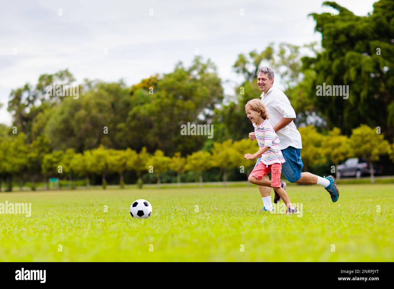 Father and son play football on outdoor pitch. Dad and kid run on park