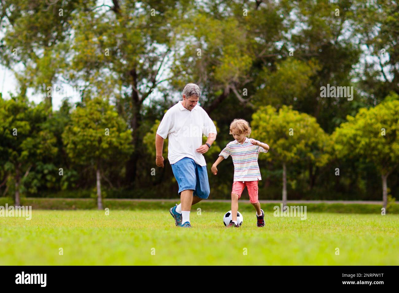 Father and son play football on outdoor pitch. Dad and kid run on park