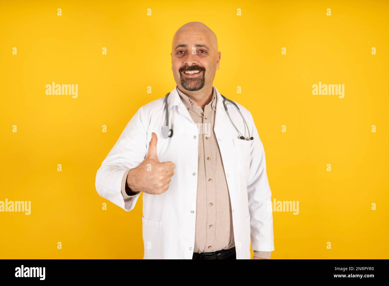 Portrait of caucasian male doctor showing thumb up gesture. Standing over warm orange studio background. Successful treatment concept idea. Copy space Stock Photo