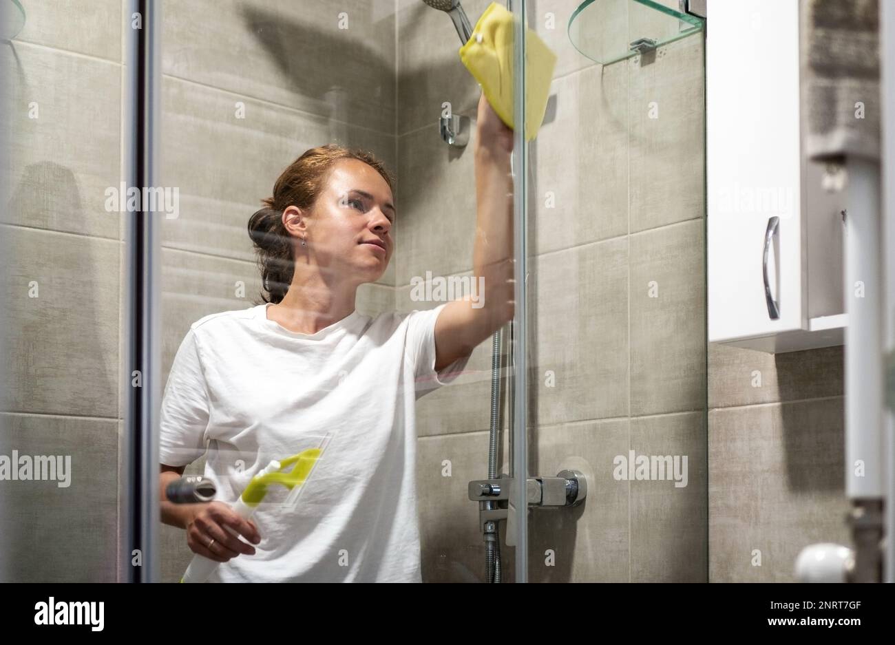Young woman cleaning glass wall in shower room. Stock Photo