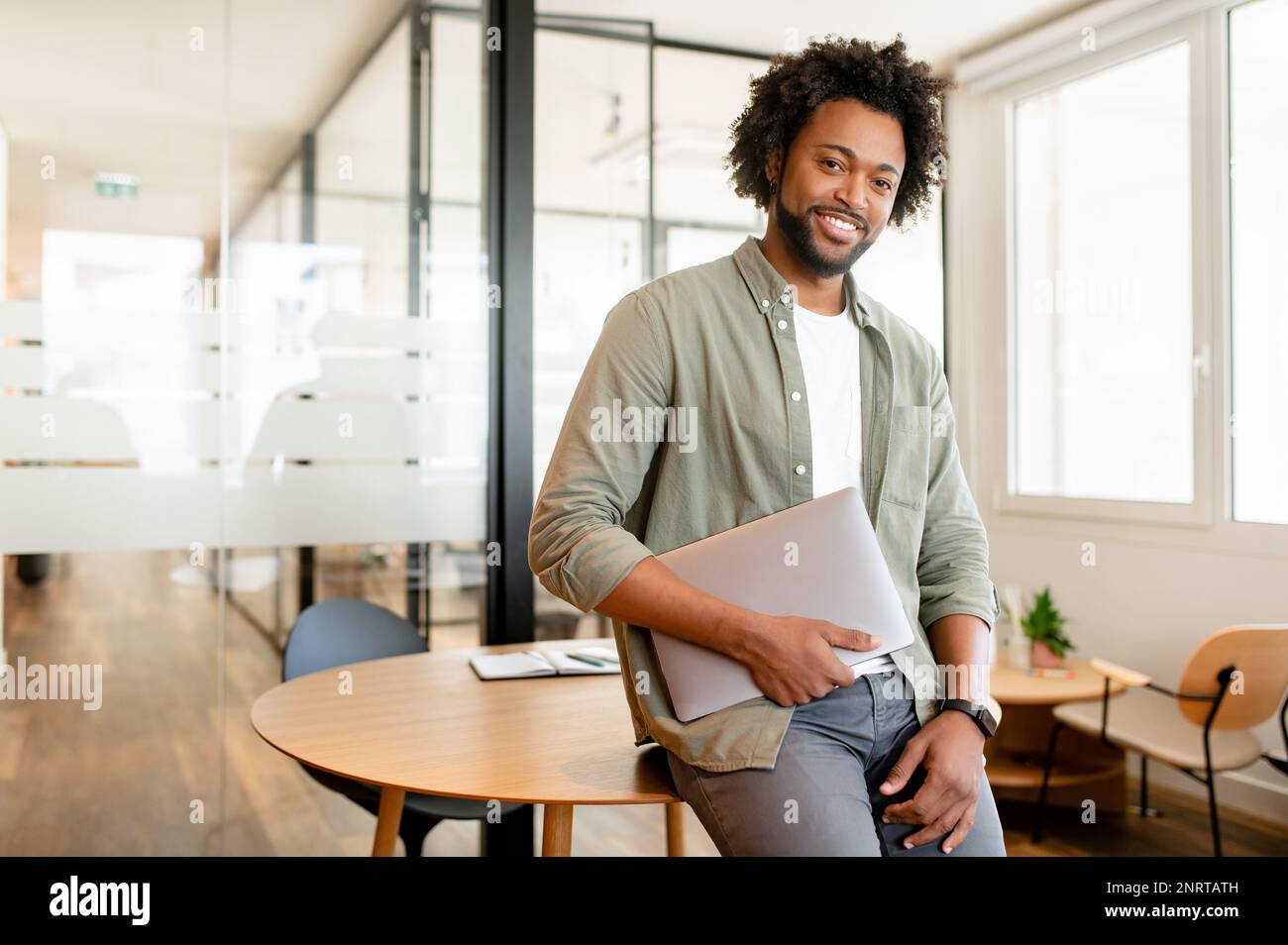 Portrait of confident African-American businessman standing with laptop, male office employee in smart casual wear looking at the camera and smiles, leaned back at te desk, ceo, manager, sturtup owner Stock Photo