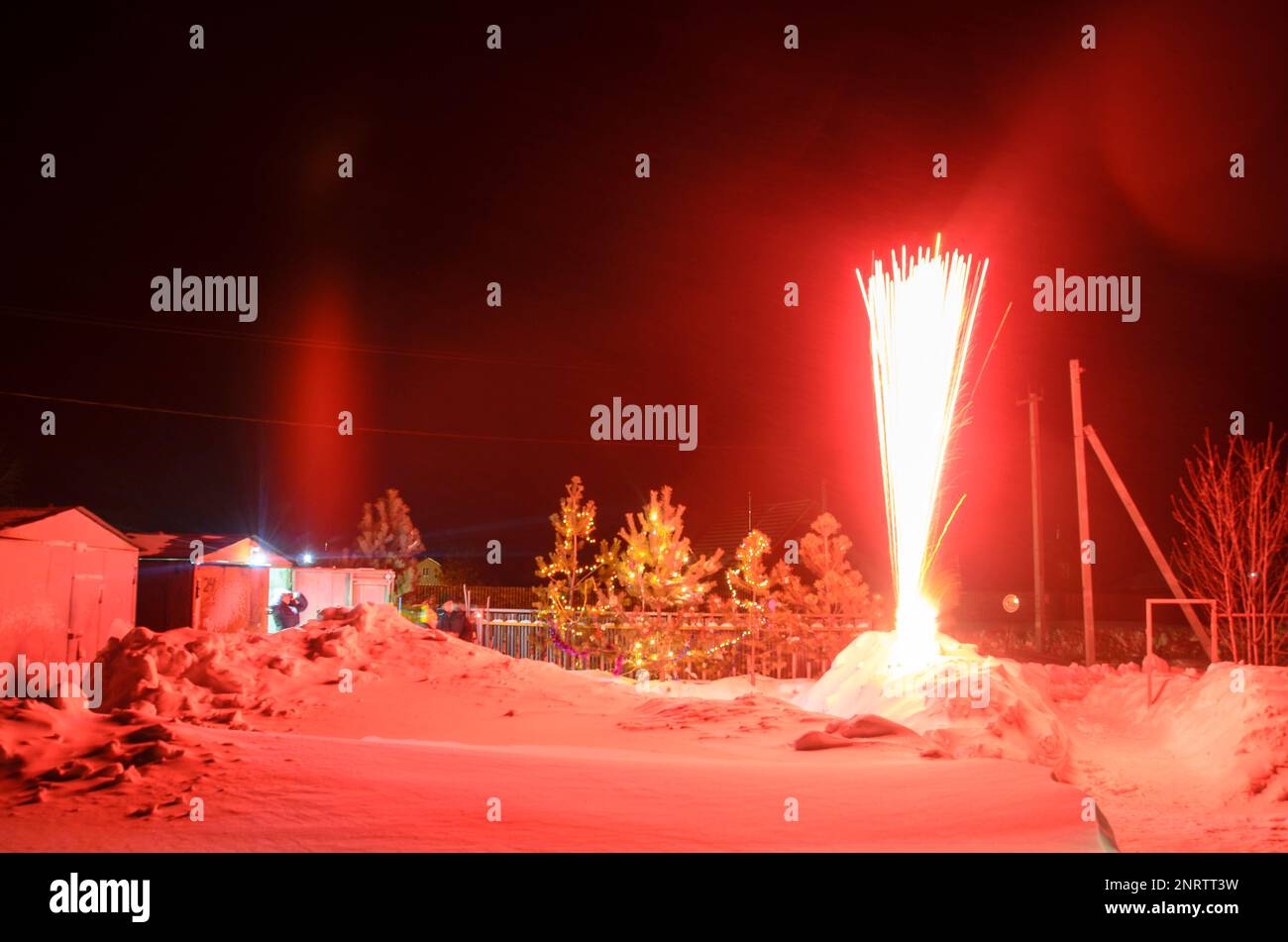 Bright flashes of fireworks in a garden plot in Siberia in winter with Christmas trees decorated with garlands on the snow at night. Stock Photo