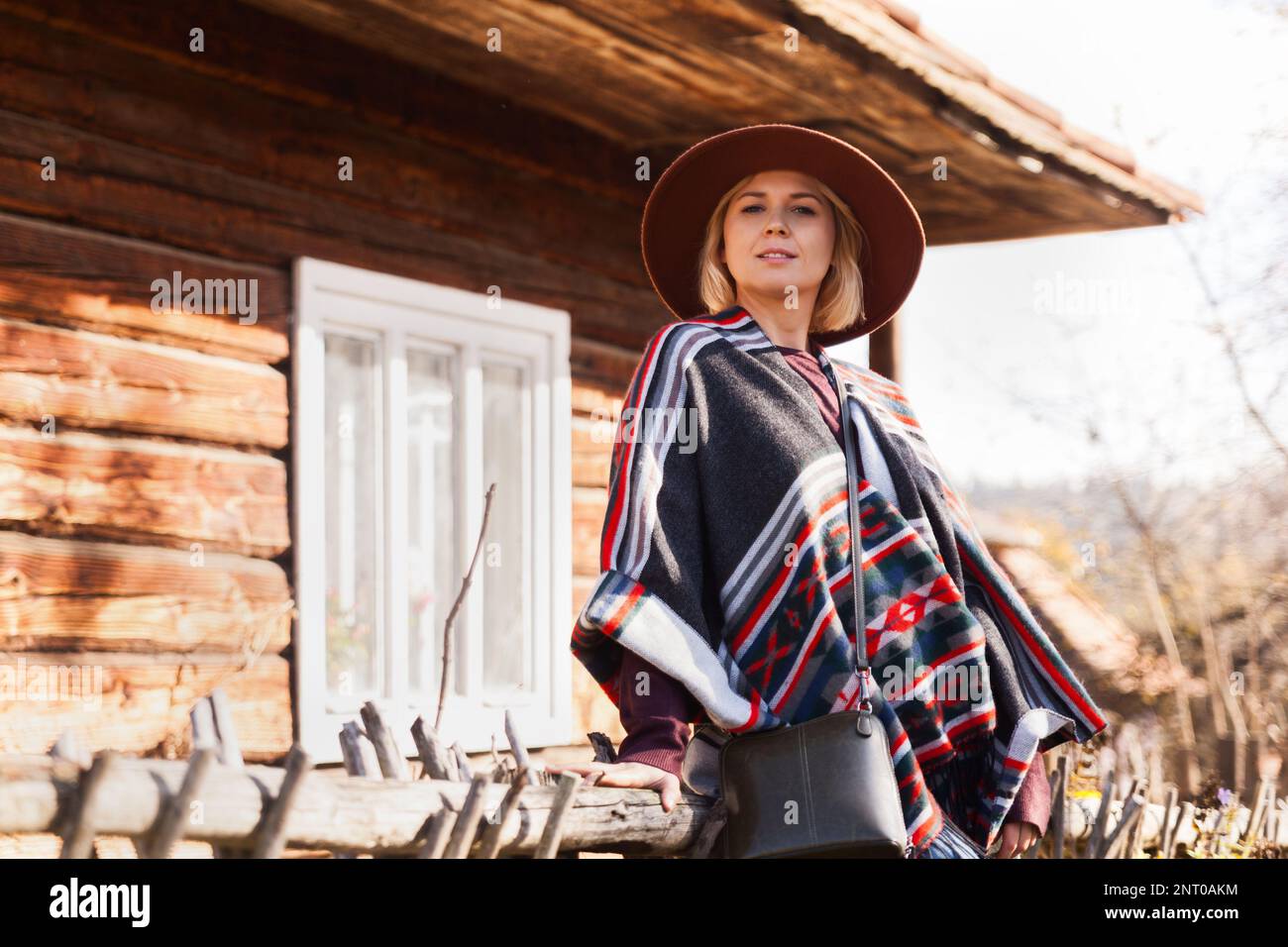 Stylish hipster traveling woman wearing  authentic boho chic style poncho and hat near old wooden countryside buildings Woman exploring nature in autu Stock Photo