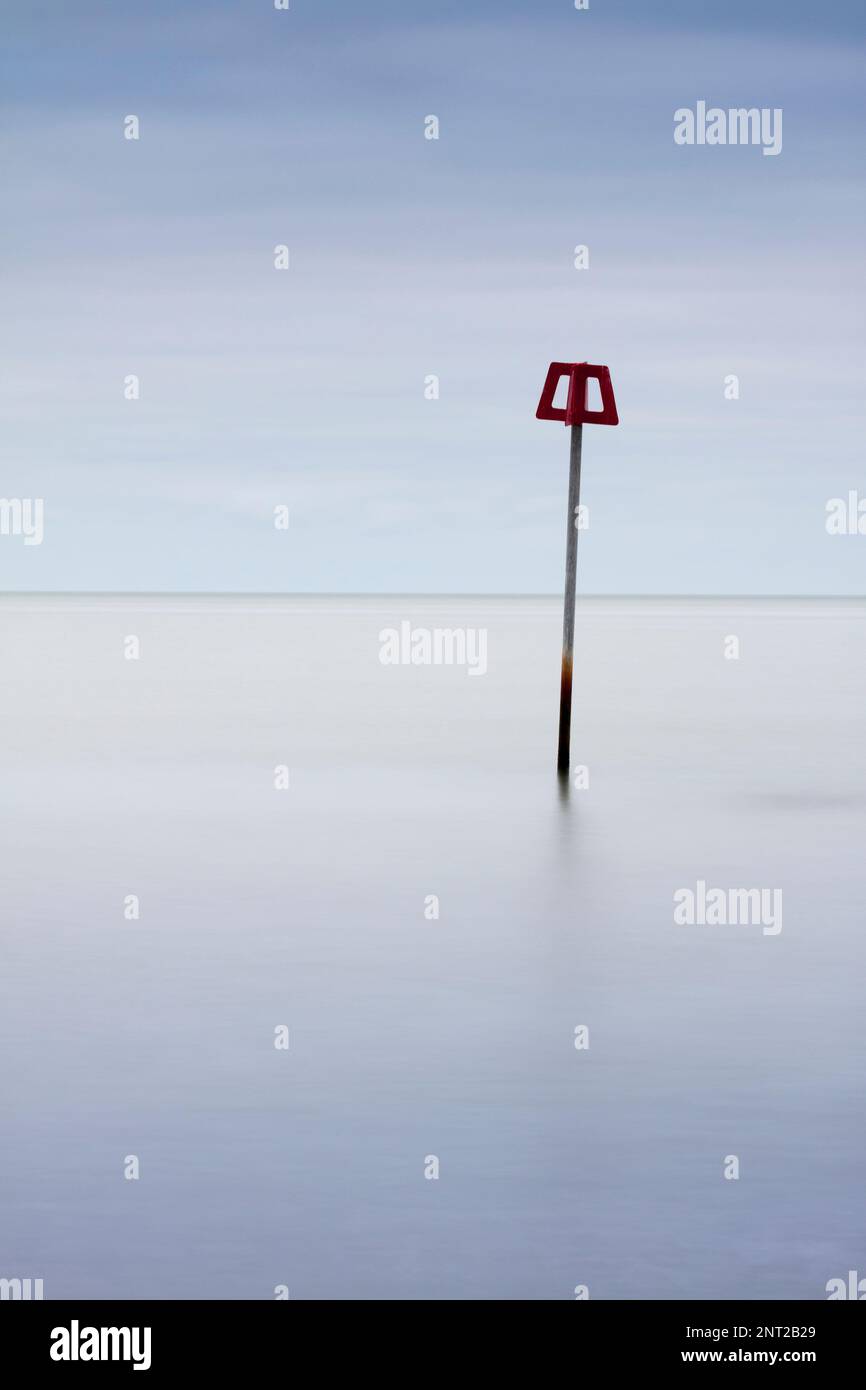A lone red beach defence groin marker amidst a perfectly calm sea and sky (portrait) Stock Photo