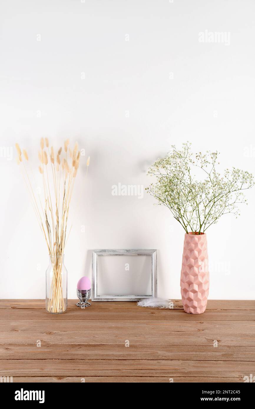 Blank frame, vases with flowers and Easter egg on table near white wall ...