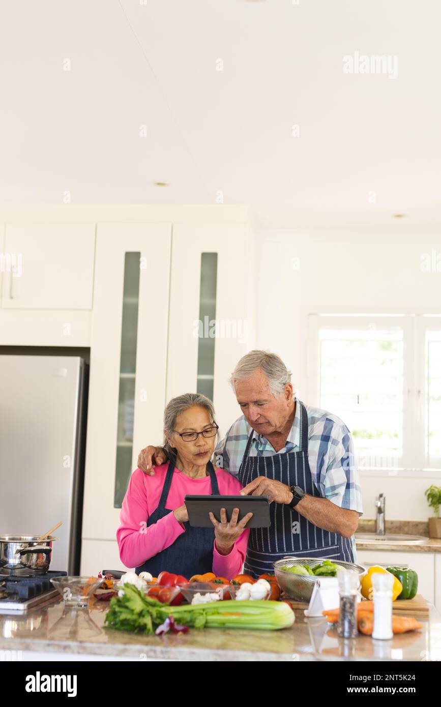 Happy senior diverse couple cooking dinner in kitchen, using tablet Stock Photo