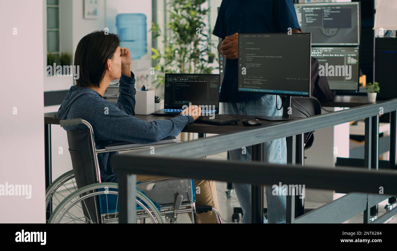 App developer talking to coder with chronic impairment in agency office, explaining source code programming on computer. Wheelchair user coding script with new user interface. Stock Photo