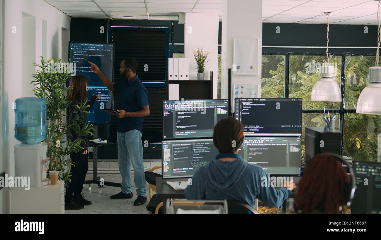 Diverse team of software engineers analyzing code on wall screen tv, using digital tablet and working with artficial intelligence. Coders parsing cloud computing project with big data. Stock Photo