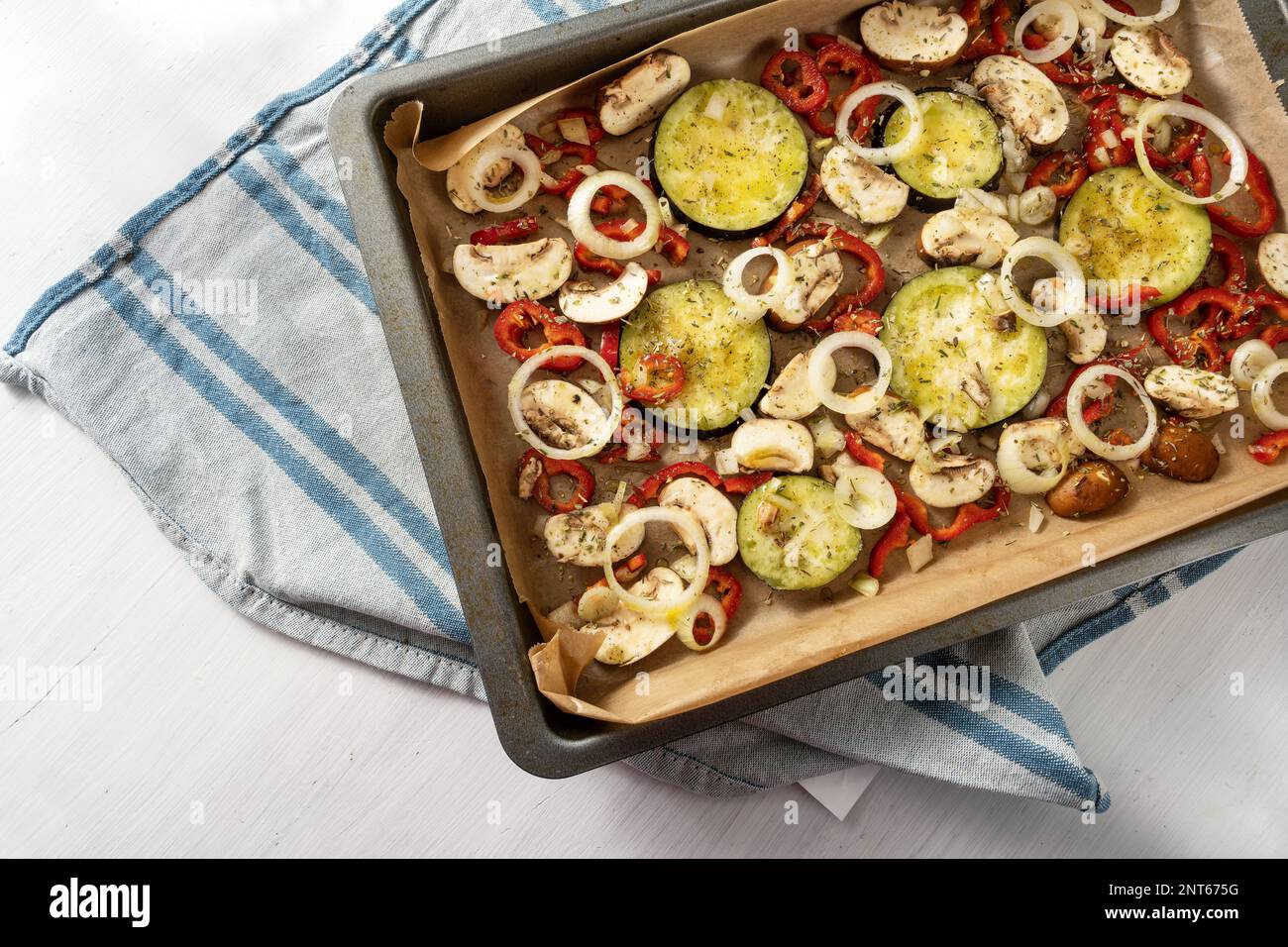 Vegetables with herbs and spices on a baking tray, blue towel and white table, Mediterranean vegetarian appetizer, copy space, high angle view from ab Stock Photo