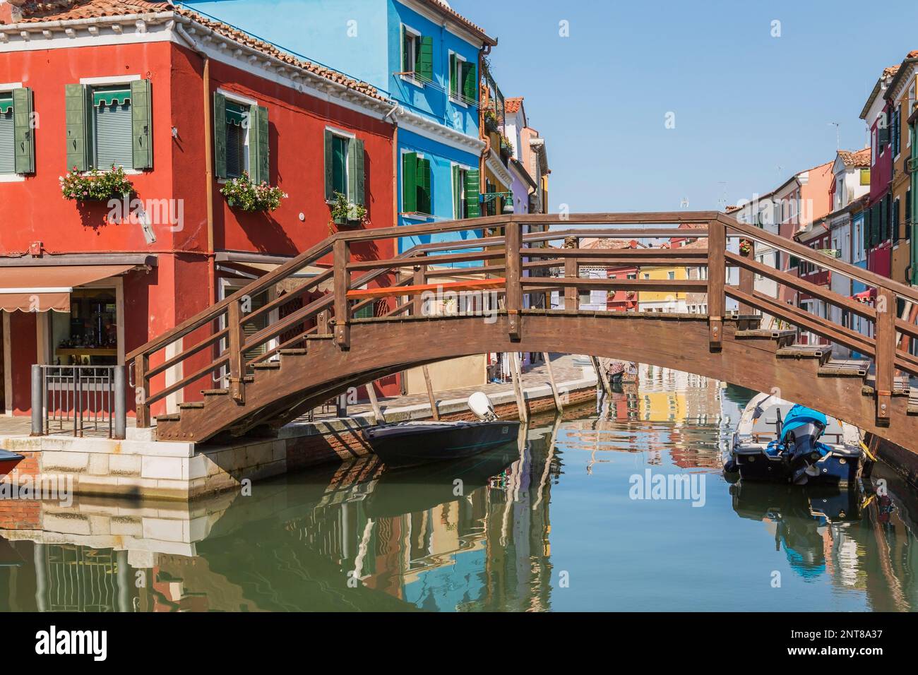 Brown wooden footbridge over canal with moored boats, red and blue stucco houses and storefronts, Burano Island, Venetian Lagoon, Venice, Italy. Stock Photo