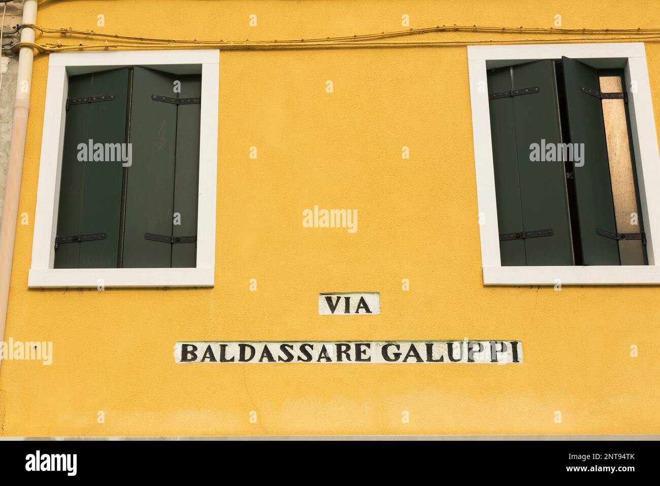 Via Baldassare Galuppi inscription on yellow stucco exterior house wall with green wooden shutters, Piazza Baldassare Galuppi, Burano Island, Italy. Stock Photo