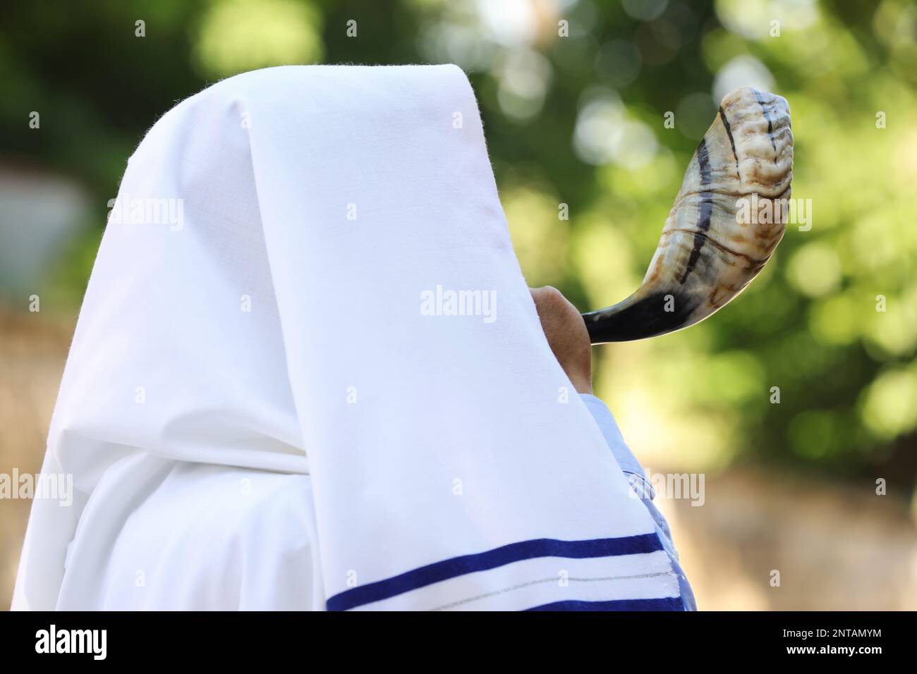 Jewish man in tallit blowing shofar outdoors. Rosh Hashanah celebration