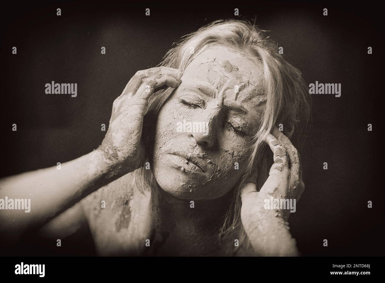 rinkled face of woman covered in dry cracked clay mask holding her head in pain, creative filtered grainy image Stock Photo