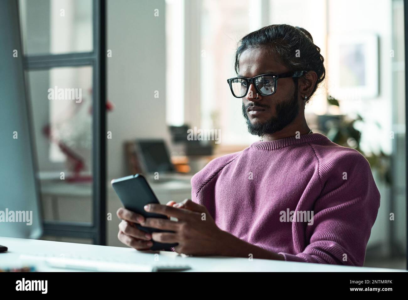 Young programmer in eyeglasses using mobile phone in his online work at table Stock Photo