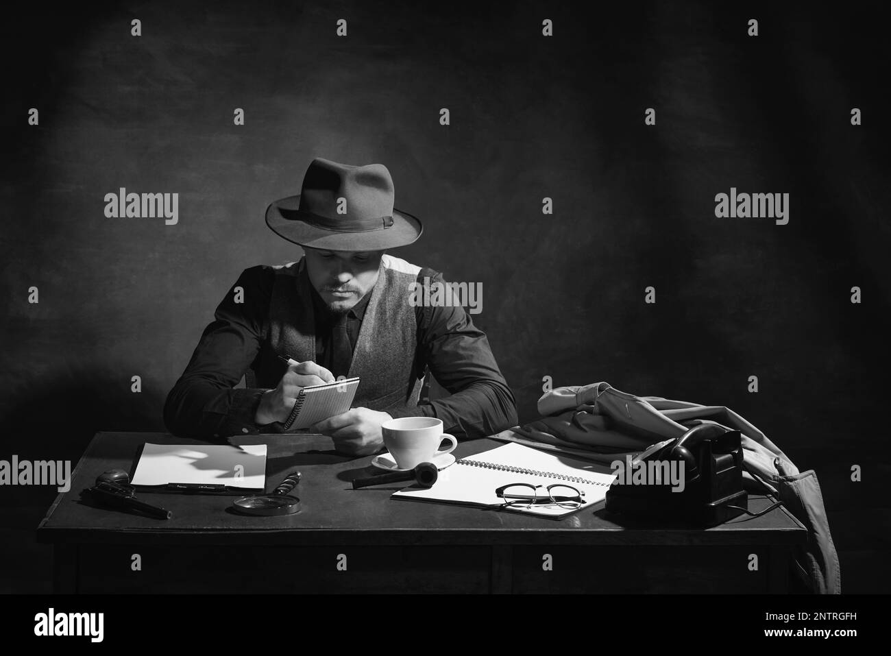 Black and white, noir photography. Serious man, professional detective in hat sitting at table and making crime notes. Investigation Stock Photo