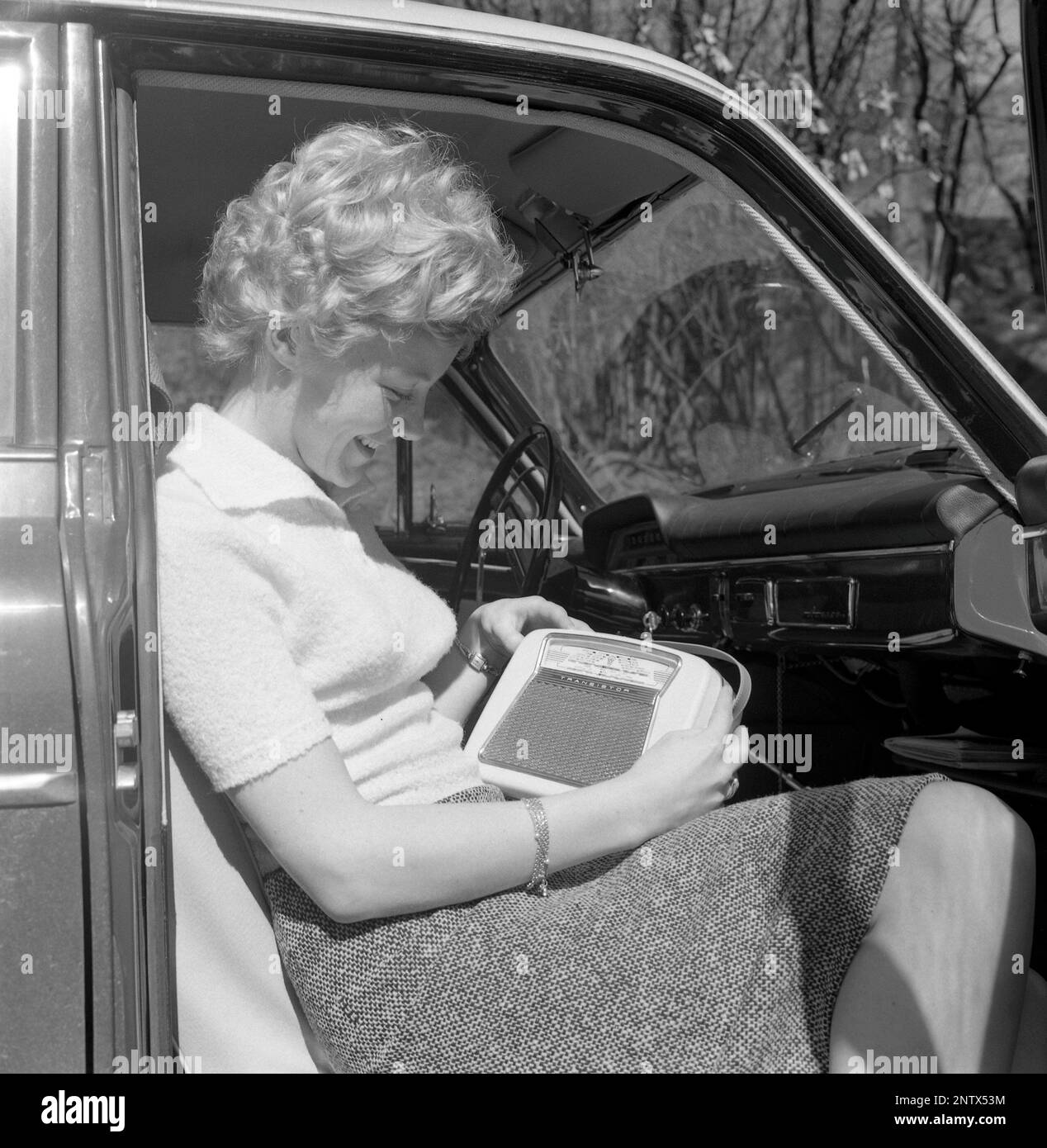 In the 1960s. A young woman in a car listening to a portable radio. Battery operated transistor radio with AM and FM bands. The radiostations and frequenses are displayed on the front, and a knob to turn and adjust to get best possible reception. Sweden 1960 Conard ref 4220 Stock Photo