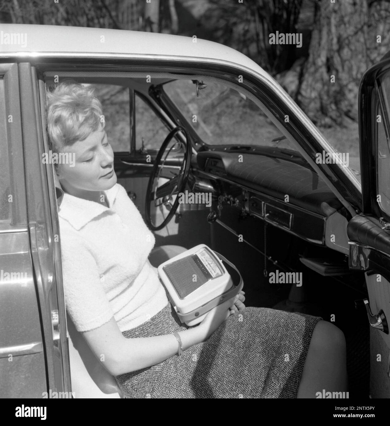 In the 1960s. A young woman in a car listening to a portable radio. Battery operated transistor radio with AM and FM bands. The radiostations and frequenses are displayed on the front, and a knob to turn and adjust to get best possible reception. Sweden 1960 Conard ref 4220 Stock Photo