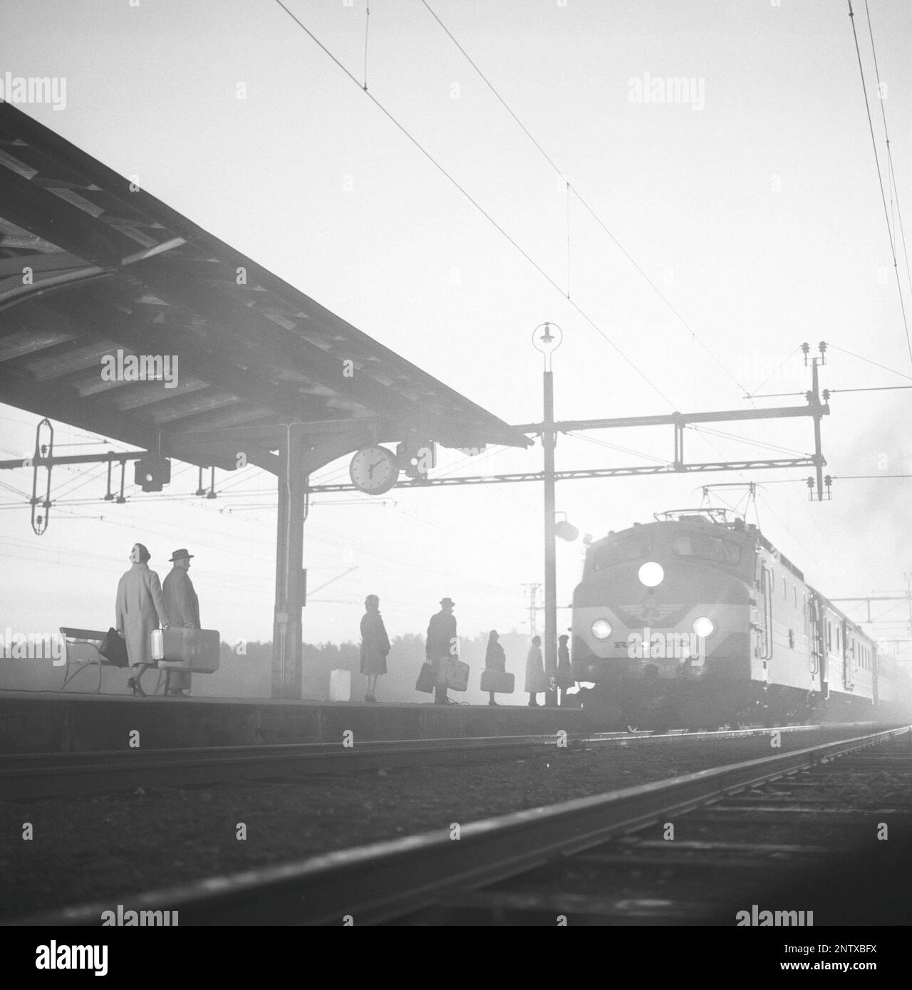 Railway station in the 1960s. Passengers are seen on the platform ready to board the train that is arriving in the misty light at ten past six. Sweden 1966 Kristoffersson ref DD106-9 Stock Photo