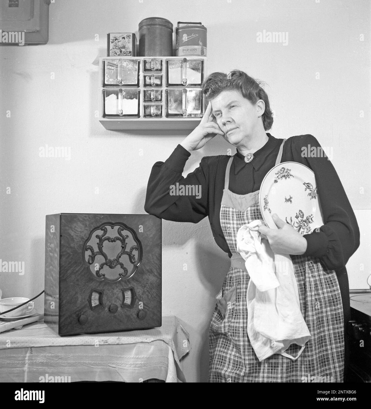 Radio listeners in the past. A woman in the kitchen has stopped what she is doing there and is seen standing listening to something important or a nice tune from the radio. On the front of the radio is a scale and when you turned the dial next to it so that the pointer was in the middle of a station name, that station was there. In addition to FM broadcasts, they also listened to foreign radio on longwave. Sweden in 1945 Kristofferson ref M4-3 Stock Photo