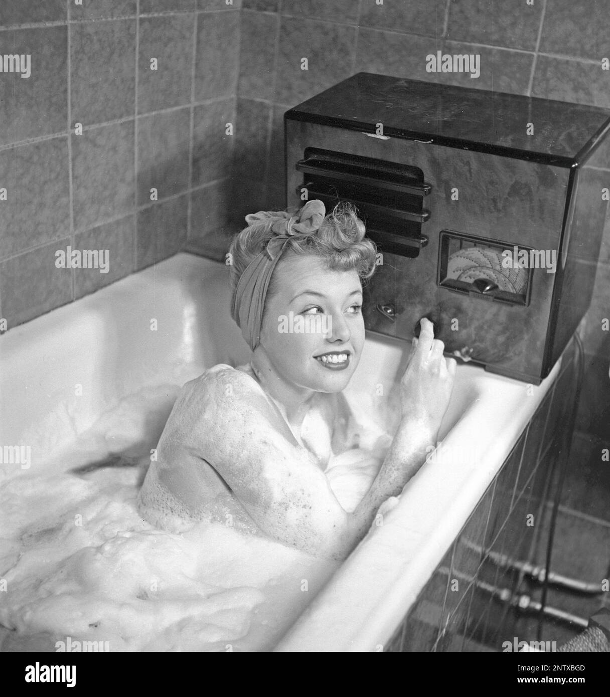 Radio listeners in the past. A young woman in the bathtub listeing to the radio that sits on the edge of the tub. On the front of the radio is a scale and when you turned the dial next to it so that the pointer was in the middle of a station name, that station was there. In addition to FM broadcasts, they also listened to foreign radio on longwave. Sweden in 1945 Kristofferson ref L180-4 Stock Photo