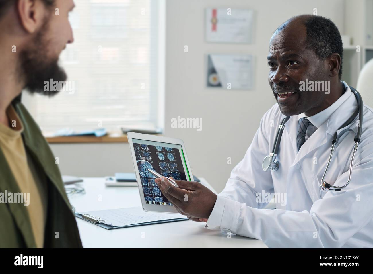 Confident mature radiologist explaining results of medical examination to young patient while pointing at brain scan on tablet screen Stock Photo