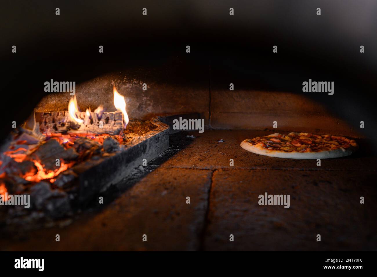 Pizza maker pushes the pizza on a wooden board into the stone oven. Burning firewood can be seen. Stock Photo