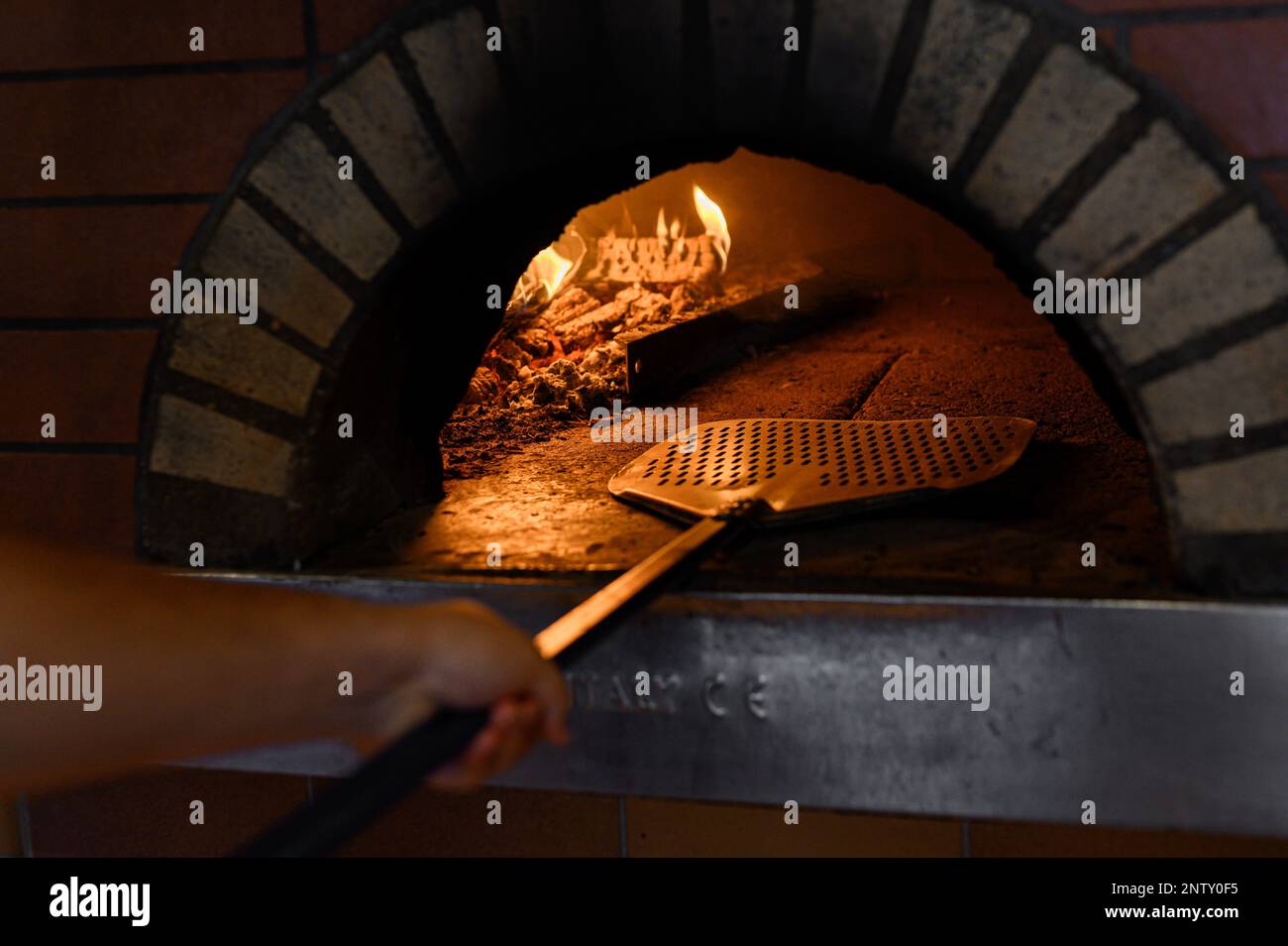 Pizza maker pushes the pizza on a wooden board into the stone oven. Burning firewood can be seen. Stock Photo