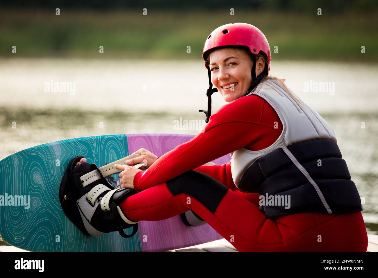 Woman in wetsuit, helmet and life vest sitting with wakeboard on a wooden pier. Sunny summer day. Safety in sport. Water sports in Finland. Insurance Stock Photo
