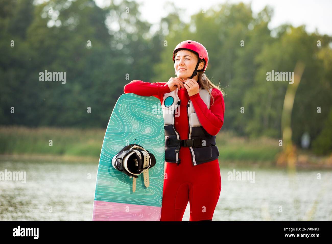 Woman in wetsuit and life vest standing with a wakeboard on a pier, fastening a helmet. Safety in sport. Water sports in Finland Stock Photo