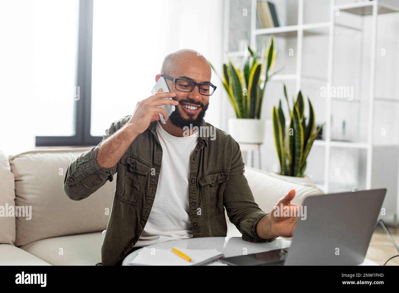 Smiling latin man using laptop and talking on cellphone, communicating with client while working online from home Stock Photo