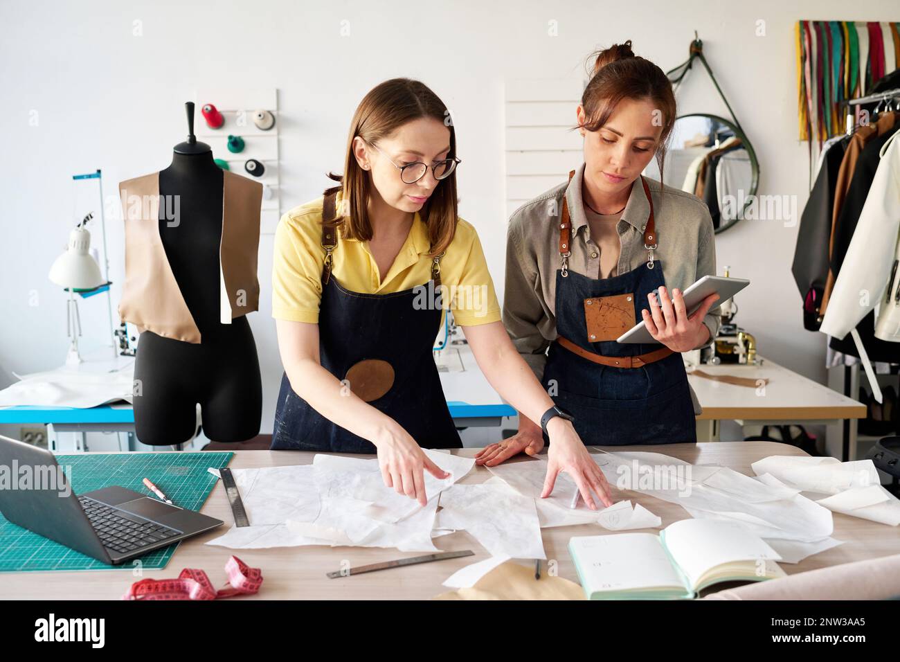 Two female tailors choosing sewing paper patterns by workplace in studio or atelier while working over new collection of leather attire Stock Photo