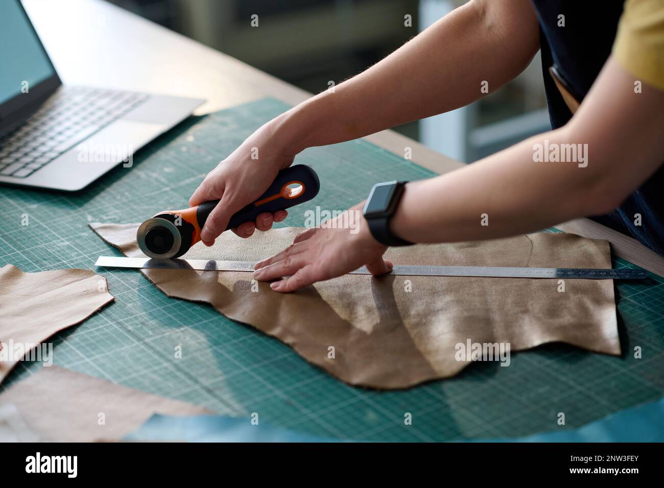 Hands of young female tailor cutting piece of beige leather on table while working over creation of new attire item in workshop or studio Stock Photo