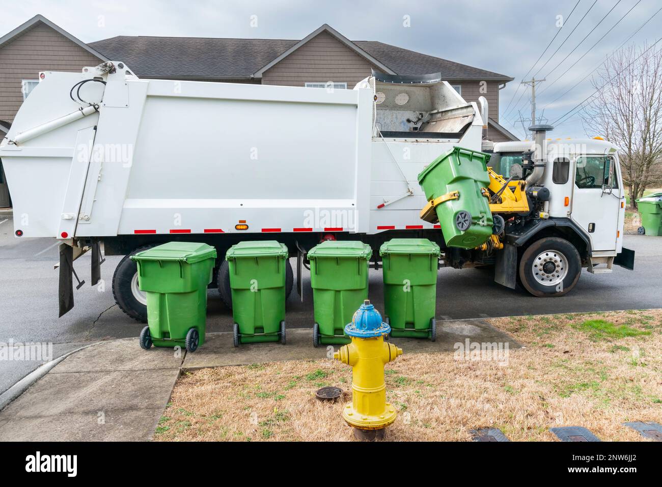 Horizontal shot of an automated garbage truck picking up trash container on curb.  Four more garbage cans are lined up waiting their turn. Stock Photo