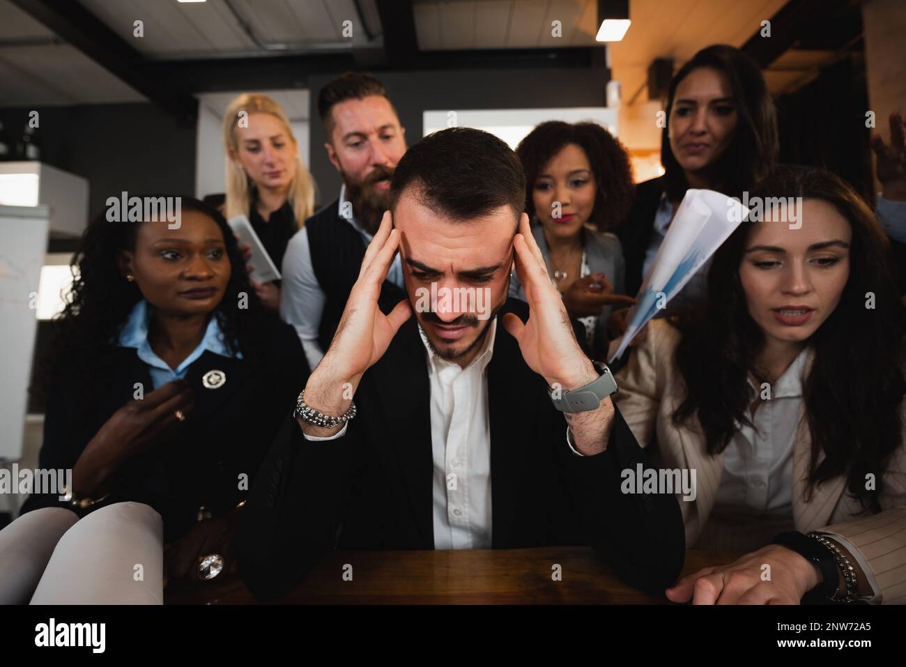 Close-up man sitting at desk with headache because colleagues are putting pressure on him. Burnout concept. Stock Photo