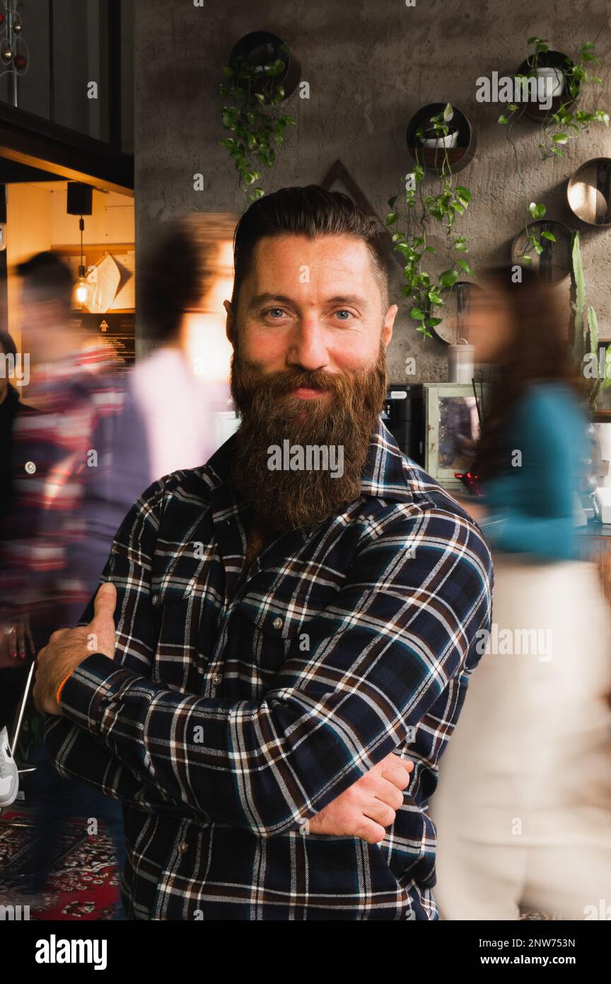 Vertical portrait of smiling man with beard and crossed arms standing looking at camera. Unrecognizable blurred people walking. Stock Photo
