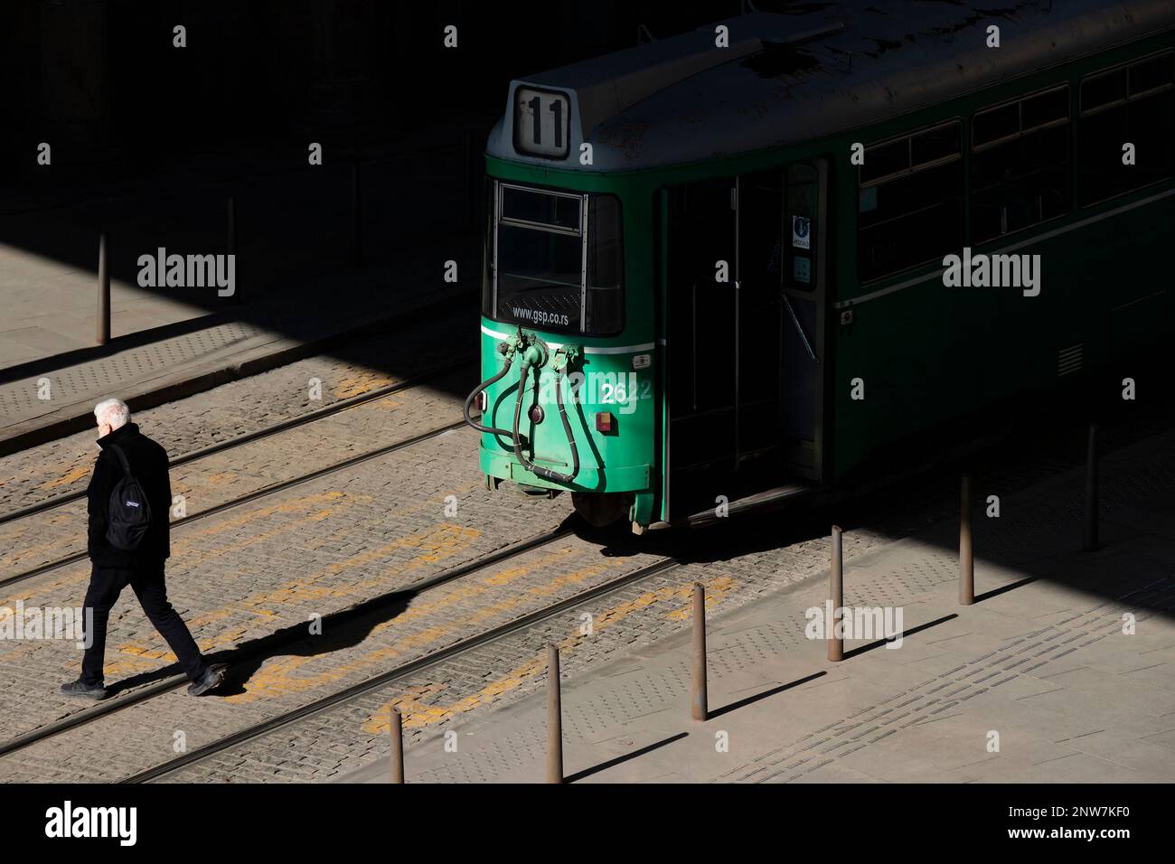 Belgrade, Serbia - February 23, 2023: Green tram number 11 standing on bus stop, and a person crossing a road behind it, high angle rear view Stock Photo