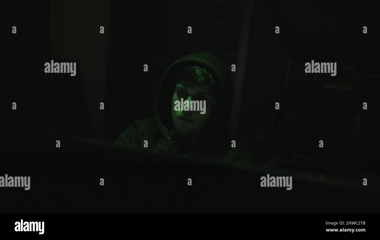 Handsome Hooded Young Computer Hacker or Programmer Working on Computer with Green Lights in Background Stock Photo