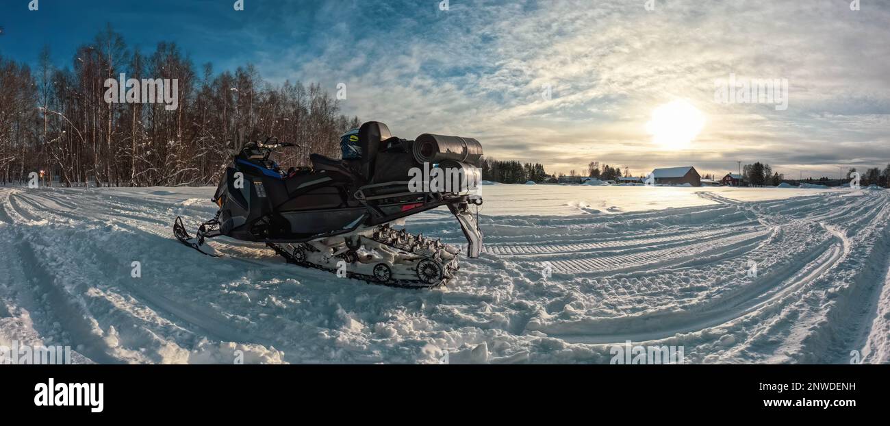 Scenic panorama of snowmobile on snow field at forest side, sunset Sun, cloudy blue sky. Side view Stock Photo