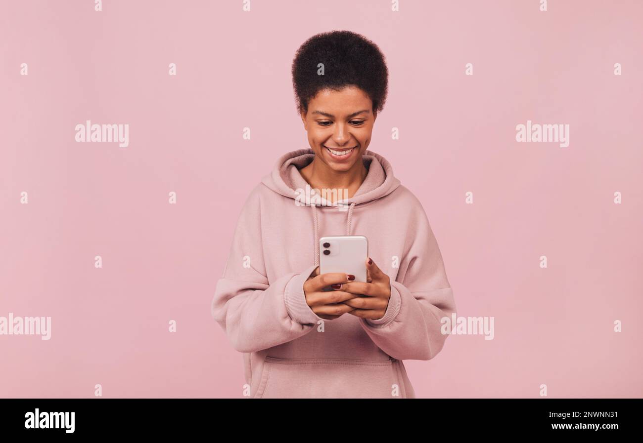 Cheerful young African woman typing message on her cell phone while standing over pink backdrop Stock Photo