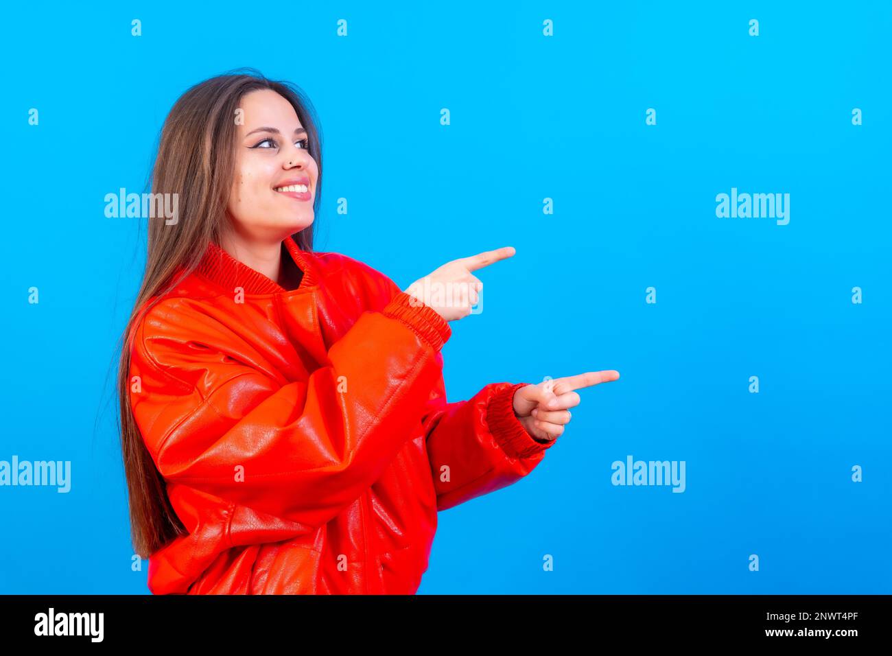 Attractive woman smiling pointing fingers at copy space on blue background, red jacket very cheerful Stock Photo