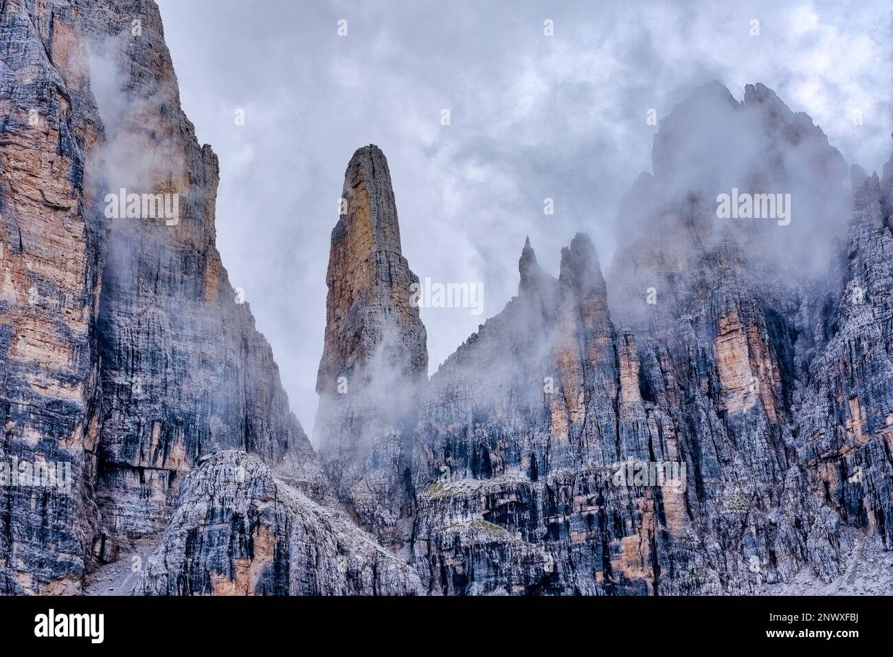 The summit of the rock needle Campanile Basso in Brenta Dolomites in the middle of surrounding rock cliffs, partially covered in clouds. Stock Photo