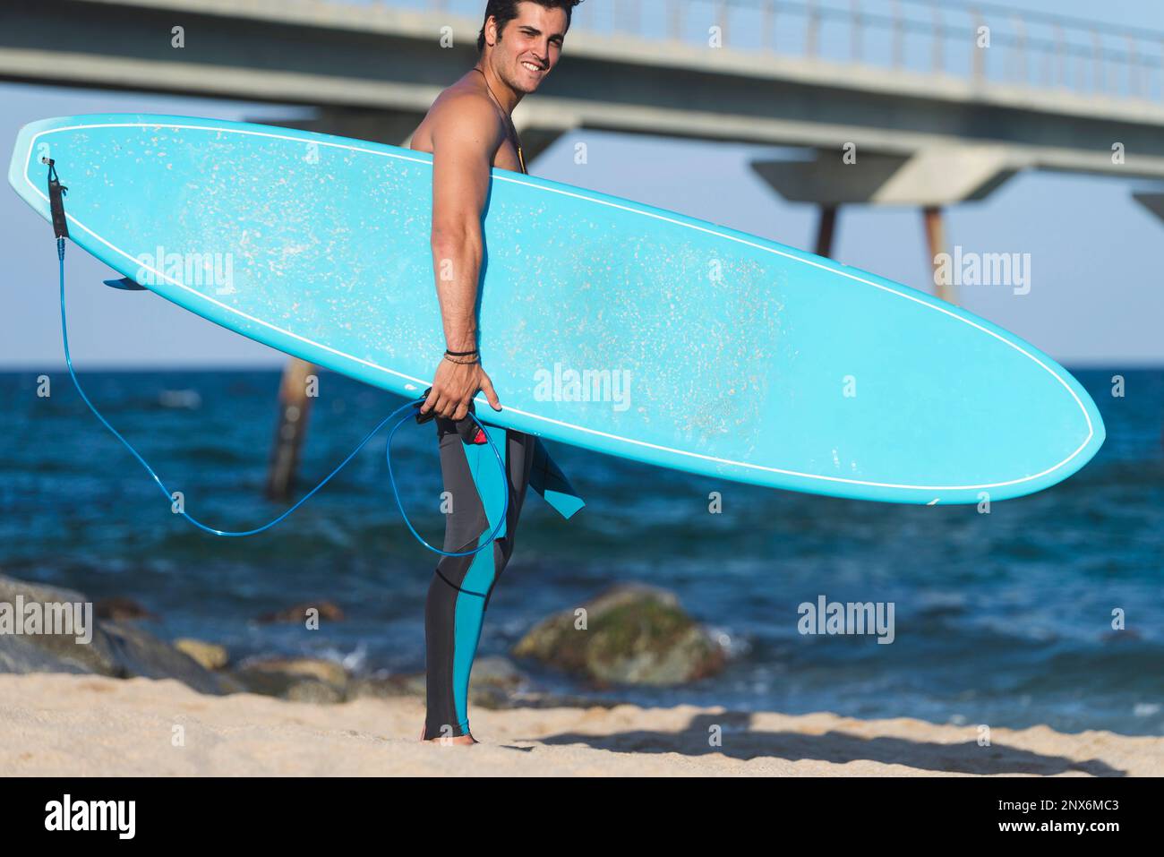 Hispanic surfer boy standing on the beach in neoprene holding his blue